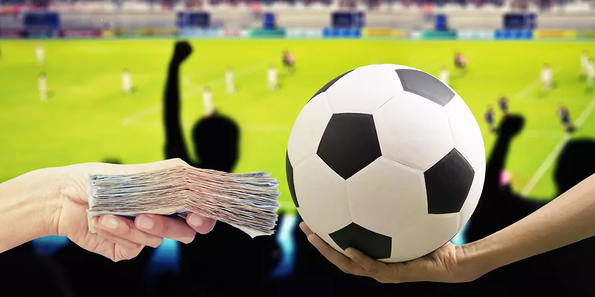 Zwei Hände tauschen Fußball gegen Geldbündel vor einem Fußballspiel im Hintergrund