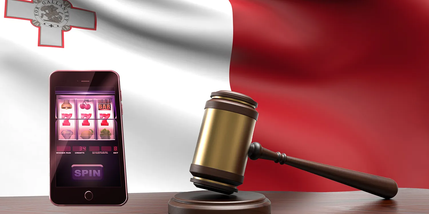 Richter-Hammer neben Smartphone, auf dem ein Online-Slot angezeigt wird, und im Hintergrund die maltesische Flagge