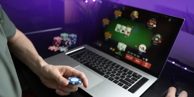 Mann am Laptop, auf dem ein Pokerspiel zu sehen ist und mit Pokerchip in der Hand
