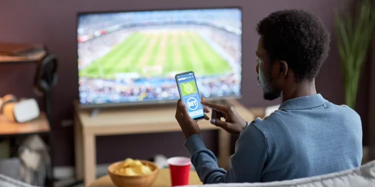 Mann vor TV mit Sportübertragung und mit Handy in der Hand, mit der eine Wette abgibt