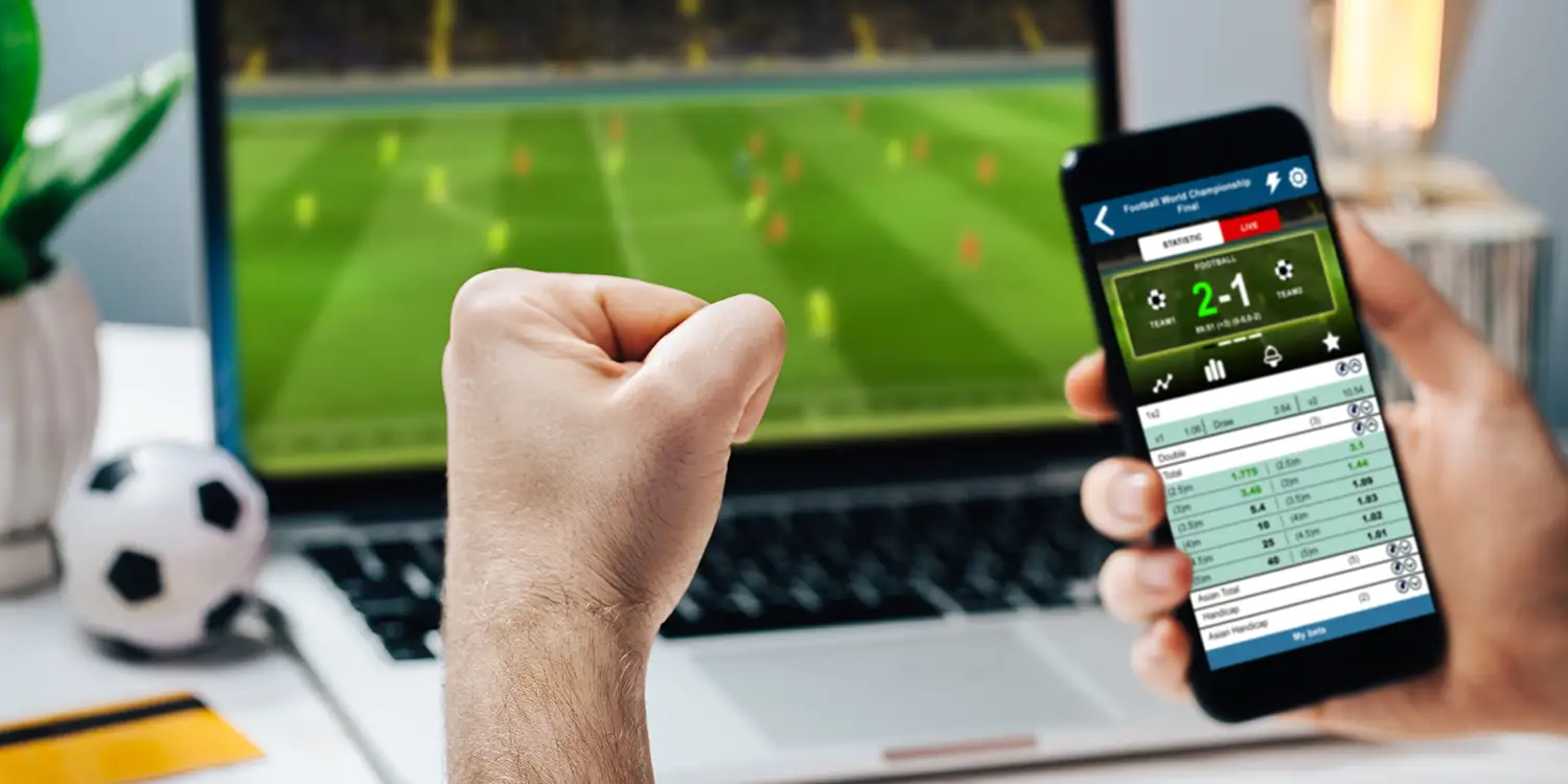 Mann tätigt Sportwetten an seinem Handy während ein Fußball-Spiel auf dem Fernseher vor ihm läuft
